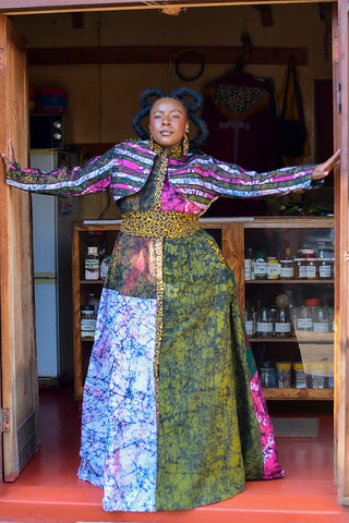 A woman stands in a doorway wearing a long, colorful patchwork dress with wide sleeves and a leopard-print waistband. The dress features multiple panels in green, blue, pink, and white patterns, with a gold leopard-print belt.