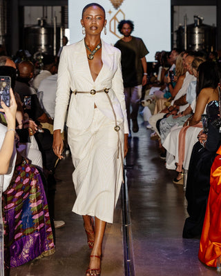 Model walking on a runway wearing a white dress with a belt, surrounded by an audience.