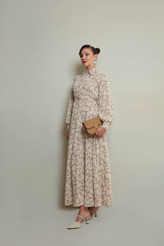 A woman modeling a floral dress with long sleeves and a round neckline, holding a clutch and wearing heels.