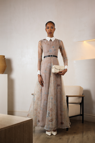 Woman in a long, light-colored dress holding a white handbag in an indoor setting.