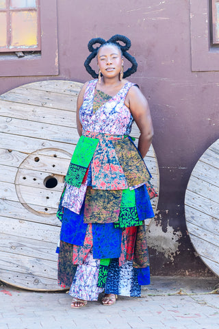 A woman stands outdoors in front of a brown wall and large wooden spool props, wearing a sleeveless tiered dress made of many colorful patterned fabric panels in blue, green, pink, red, beige, and black.