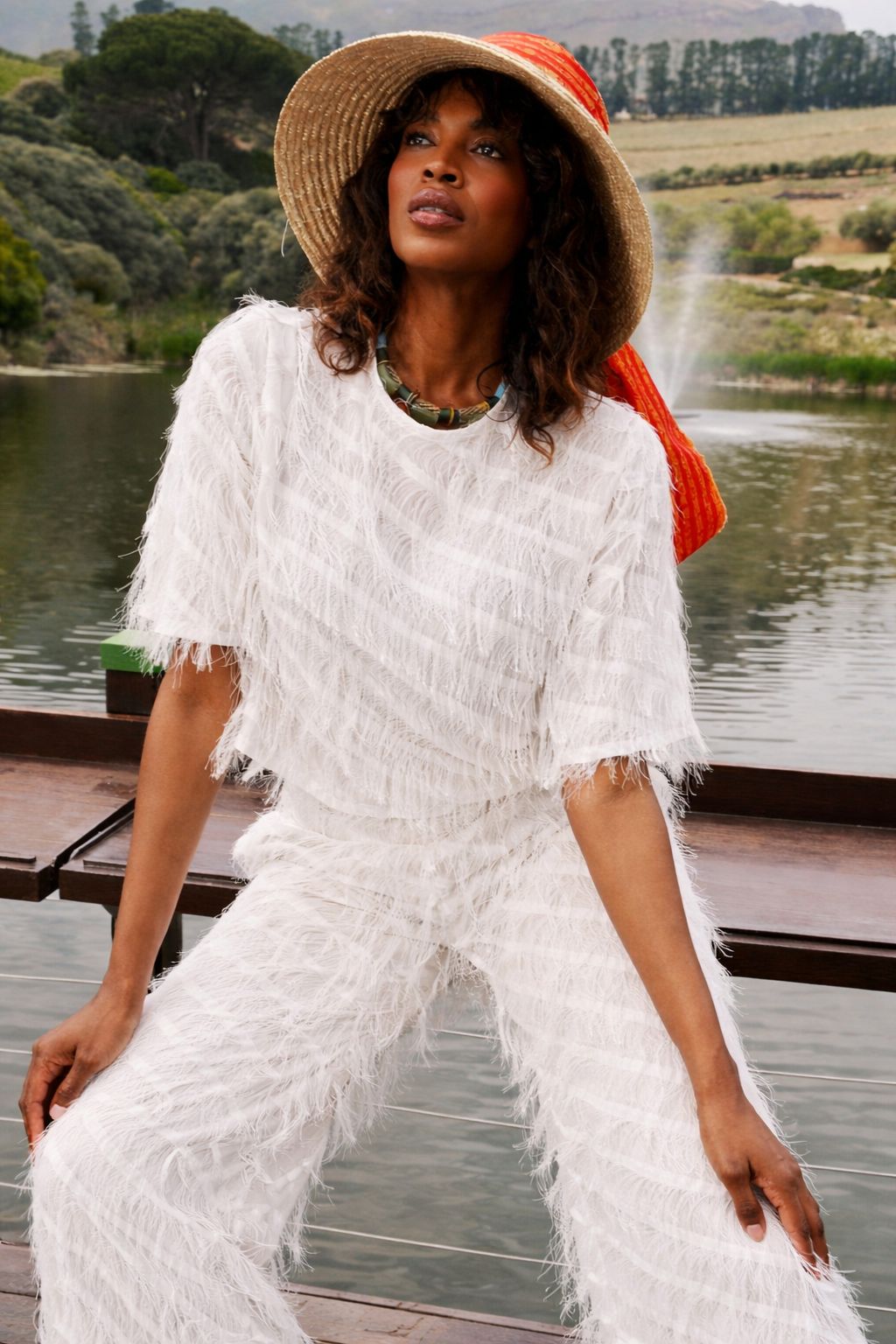 Woman in a white outfit with fringe sitting by a lake wearing the NALA Prive Chiffon Fringe Set White for House of NALA. 
