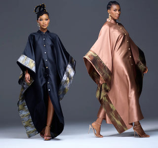 Two models wearing traditional African-inspired capes (agbadi) in black and champagne pink satin, both featuring wide patterned borders in gold, green, and gray, set against a dark gray studio background for House of NALA.