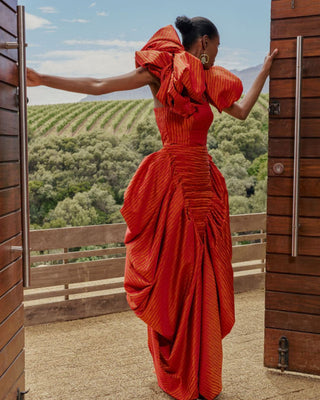 Woman in an orange evening dress standing on a wooden deck with a scenic background. By NALA Privé at House of NALA 
