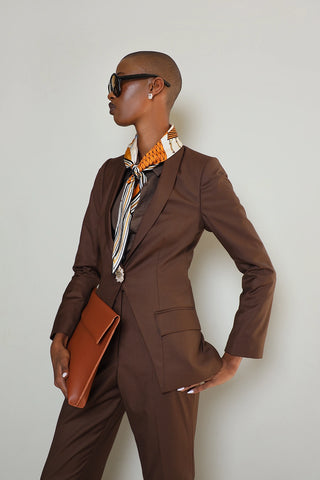 A woman modeling a brown suit jacket with a triangular bottom design and a one-button closure. She is also wearing sunglasses and carrying a clutch.