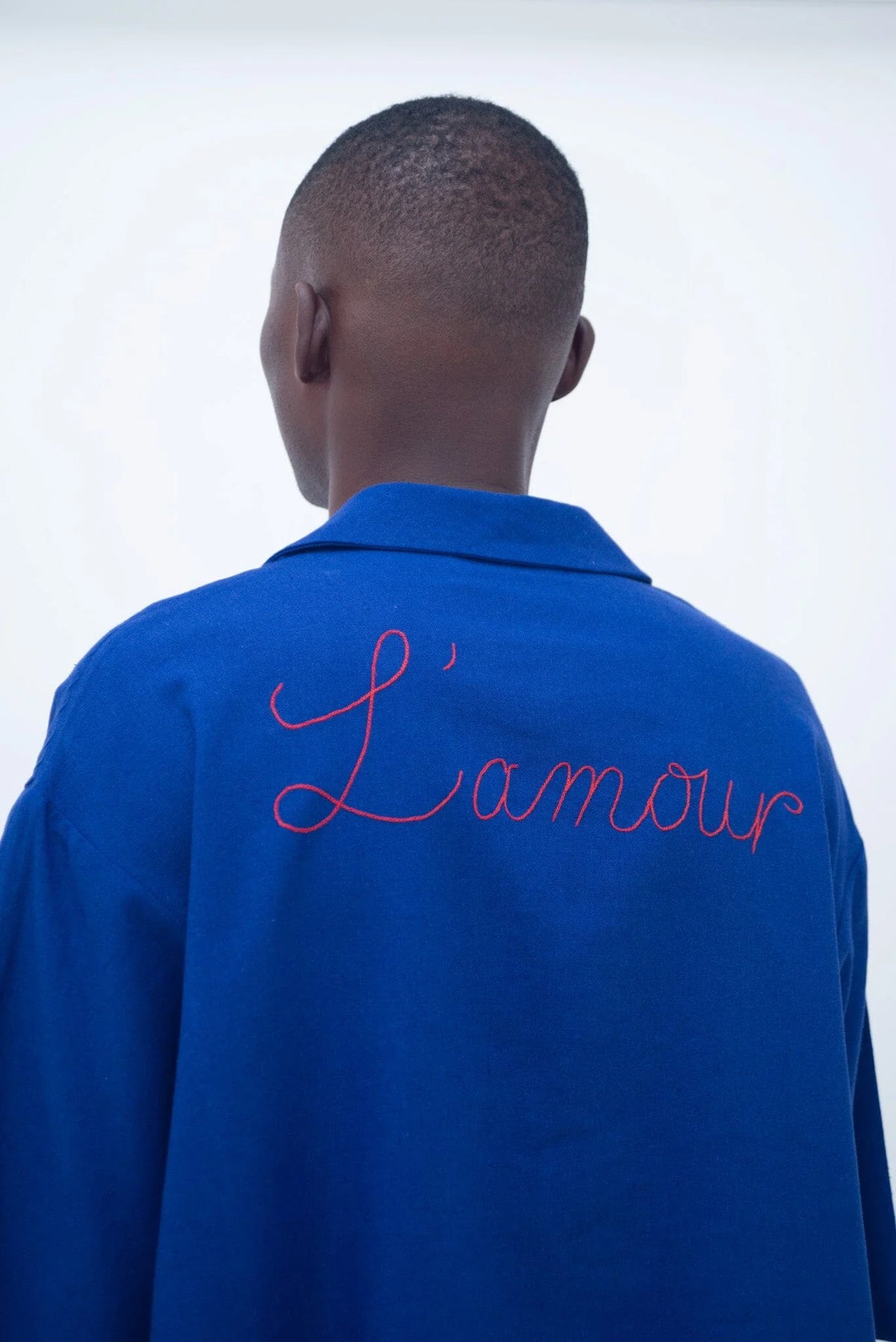 A person viewed from behind wearing a bright blue collared shirt or jacket with the word "L'amour" embroidered in red script across the upper back, against a plain light background.